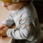Kleiner Junge in grauen Body mit senfgelben Leoparden und Pflanzen drauf. Trägt die passende senfgelbe Mütze