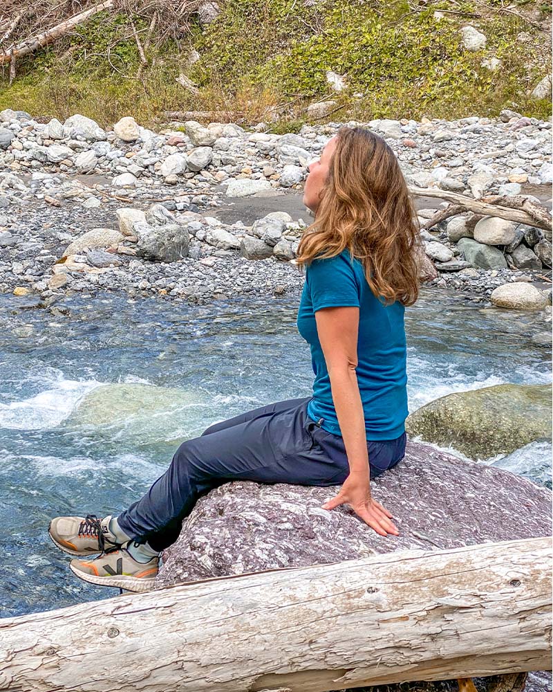 Naturbild einer Frau, die im Fluss auf einem Stein sitzt und in die Ferne blickt. Sie trägt ein türkises Tshirt von der Marke ROTAUF