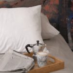 Ausschnitt eines Schlafzimmer mit weisser Bettwäsche von Storystabrics. Auf dem Bett liegt zu dem ein Tablett mit einer Zeitung, einer Bialetti und einer Tasse Kaffee