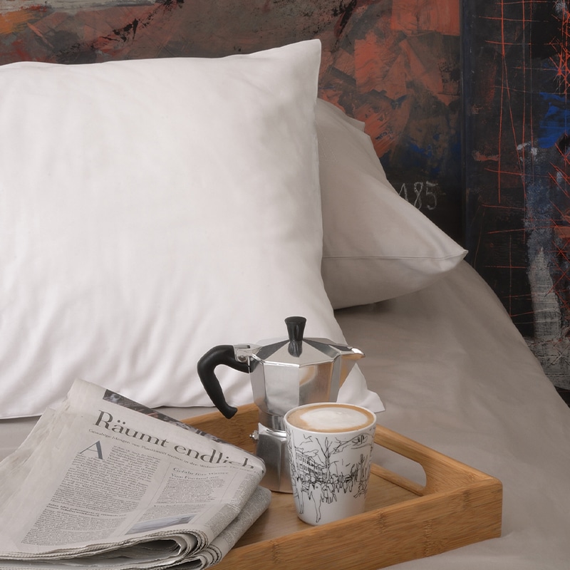 Ausschnitt eines Schlafzimmer mit weisser Bettwäsche von Storystabrics. Auf dem Bett liegt zu dem ein Tablett mit einer Zeitung, einer Bialetti und einer Tasse Kaffee