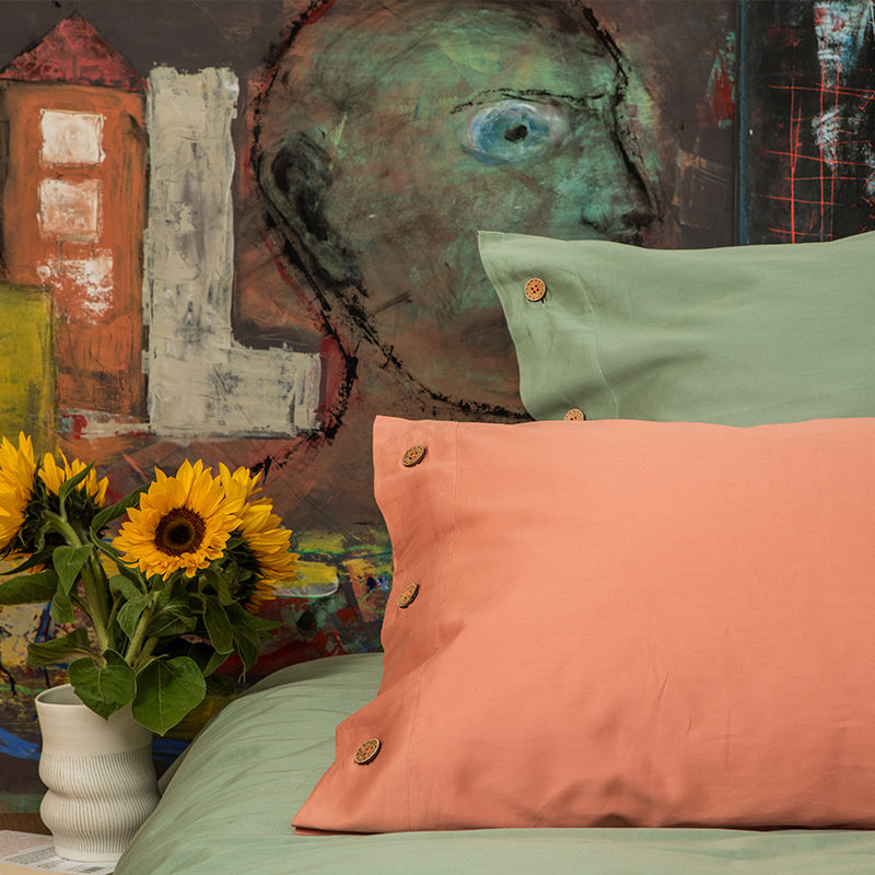 Auschnitt eines Schlafzimmers mit hellgrüner und terrakotta Bettwäsche und Blumen in einer Vase