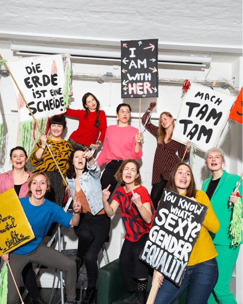 Gruppenfoto vom einhorn Frauenteam. Sie haben bunte Kleider an und halten feministische Tafeln in die Höhe. Sie stehen in einem rustikalen Raum.