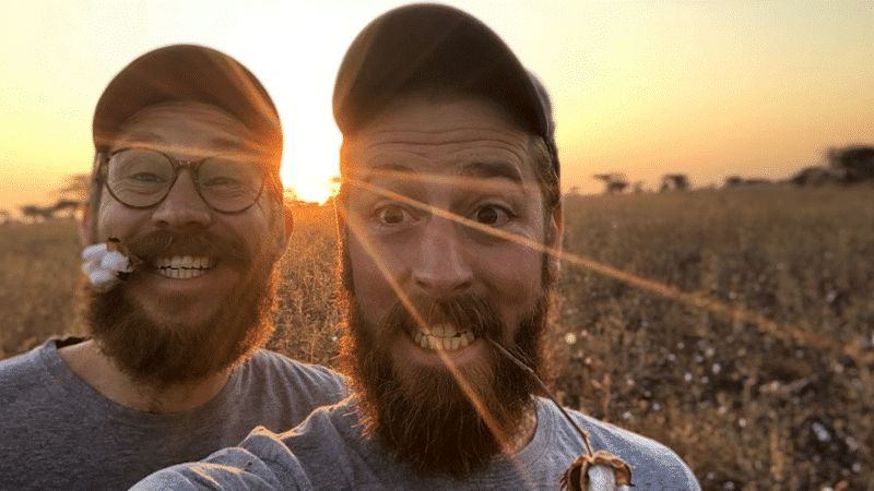 Zwei Mitarbeiter von einhorn stehen im Sonnenuntergang auf einem Baumwollfeld. Die Männer haben einen Bart, einen Sonnenhut auf und tragen beide graue Tshirts. Der linke Mann hat eine Baumwollpflanze zwischen den Zähnen.